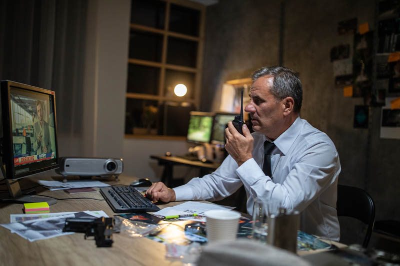 Side view of gray-haired police detective in white shirt and tie working late at night in his office, sitting at the desk, using walkie-talkie and looking at computer monitor